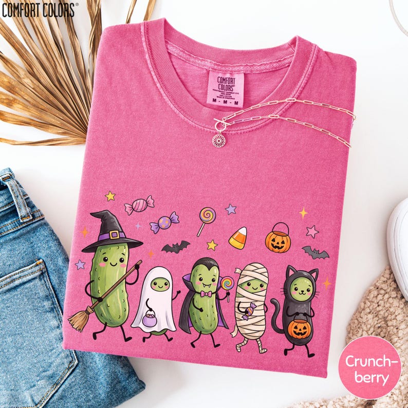 a pink t-shirt with a cartoon-style illustration of a group of anthropomorphic vegetables dressed in Halloween costumes, including a witch, pumpkin, skeleton, and ghost.
