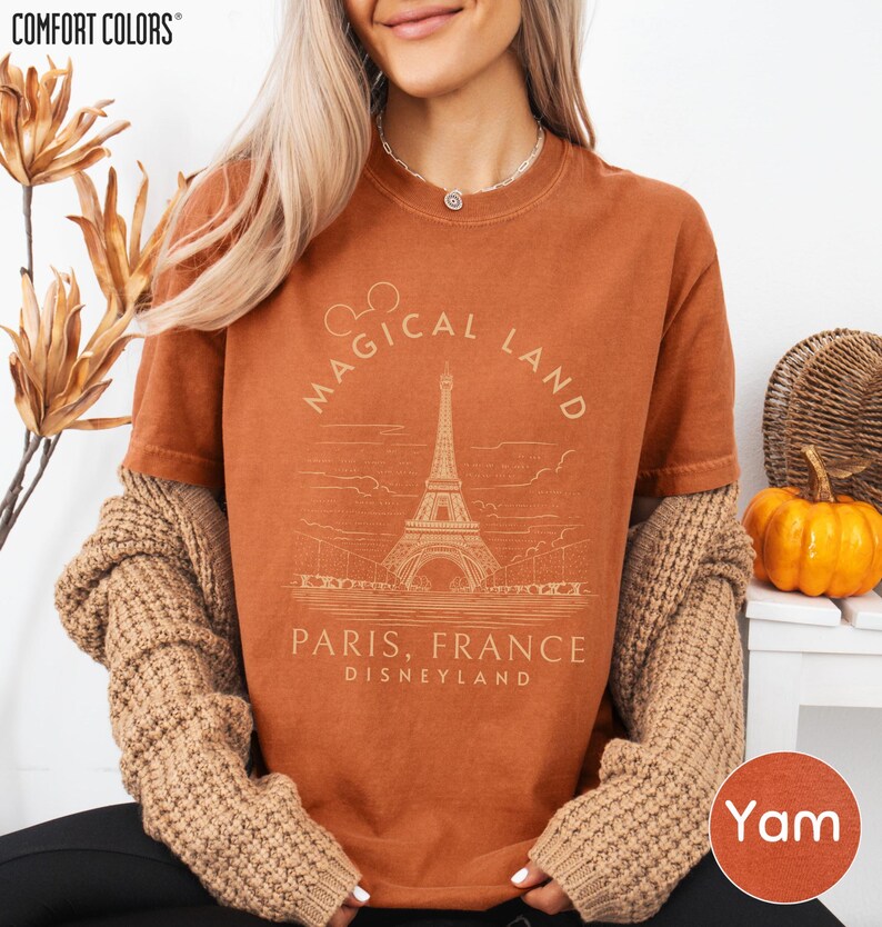 A woman wearing a brown sweater and a t-shirt with a graphic of the Eiffel Tower and text that says "Magical Land Paris, France Disneyland" is sitting in front of a plant and a pumpkin.