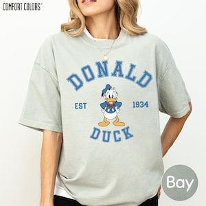 A woman wearing a grey t-shirt with a graphic of a cartoon duck and text that says "DONALD DUCK EST. 1934".