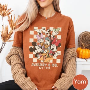A woman wearing a t-shirt with a Disney character graphic and text, sitting in front of a plant and a pumpkin.