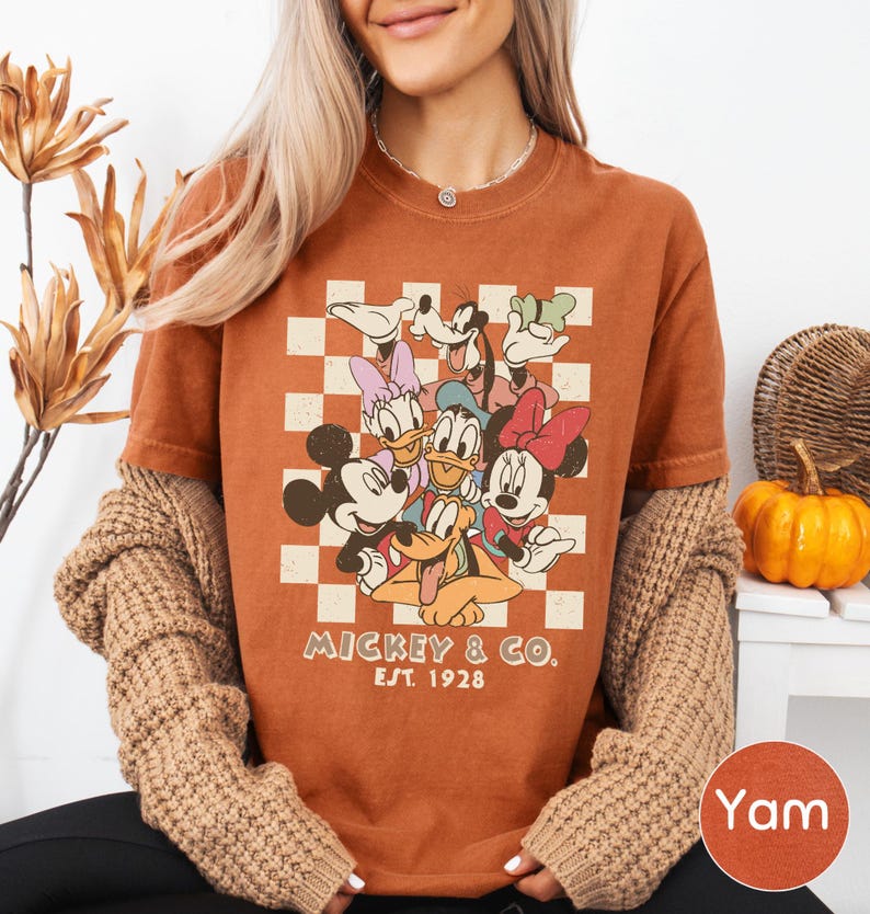A woman wearing a t-shirt with a Disney character graphic and text, sitting in front of a plant and a pumpkin.