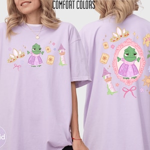 May include: Orchid-colored t-shirt with a cartoon design featuring a green frog in a pink dress, a gold crown, and a tower. The back of the shirt has a similar design inside a pink oval frame. The text "COMFORT COLORS" is at the top.