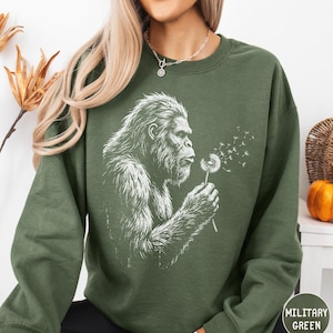 May include: A military green long-sleeve sweatshirt featuring a white graphic print of a hairy creature blowing on a dandelion, with seeds drifting into the air. The sweatshirt has a crew neckline and long sleeves, providing a simple, solid-colored base.