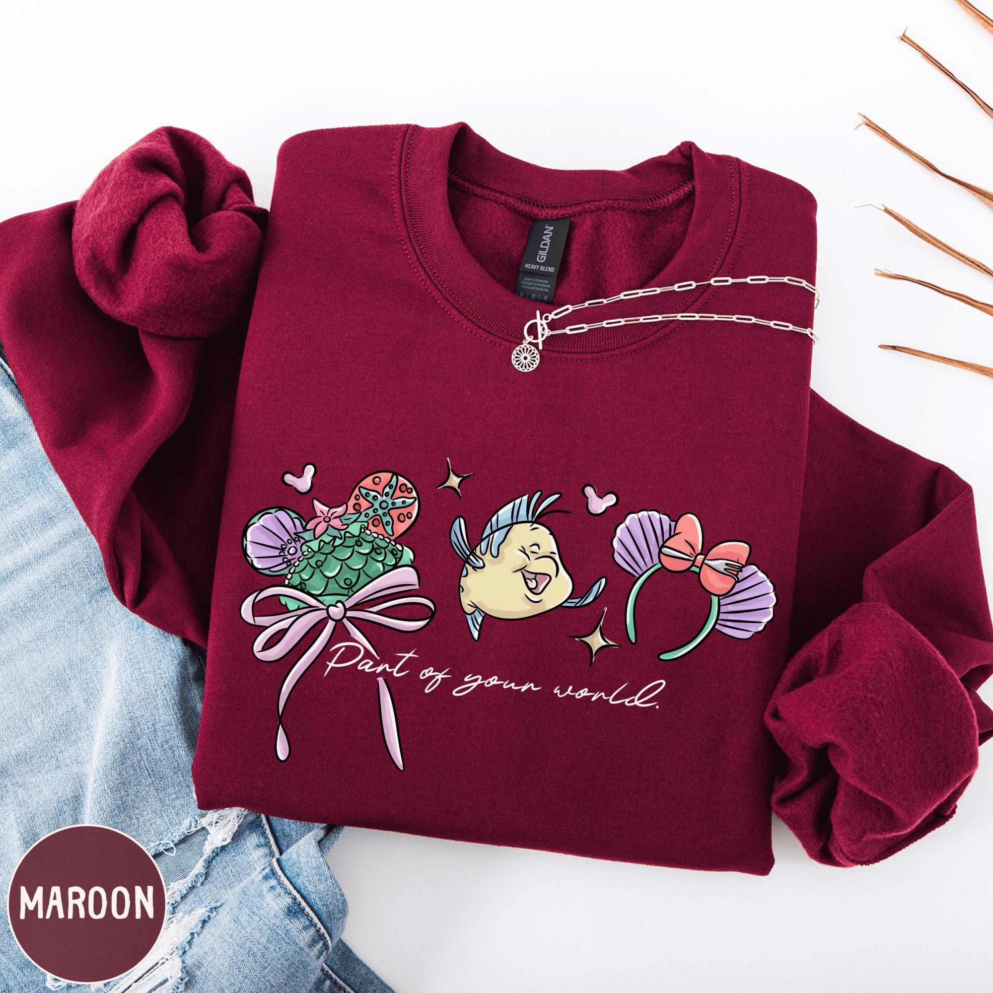 A burgundy sweatshirt with a graphic design featuring a mermaid character and the text "Part of your world" printed on it.