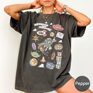 A person wearing a black t-shirt with a graphic design featuring various cartoon characters and icons, including a cowboy hat, a toy car, and a red ribbon.