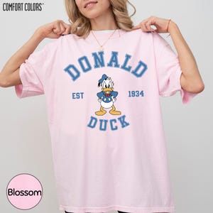 A woman wearing a pink t-shirt with a graphic of a cartoon duck and the text "DONALD DUCK EST 1934" printed on it.