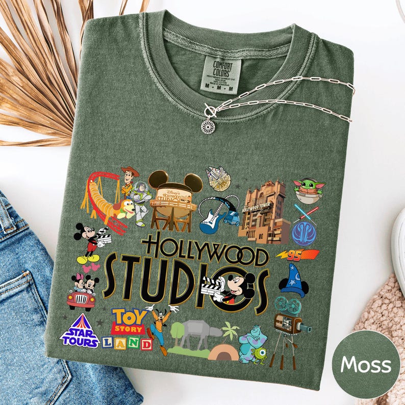 a green t-shirt with various Disney and Pixar characters and logos, including Mickey Mouse, Minnie Mouse, Woody, and others, along with the text "Hollywood STUDIOS" and "Toy Story Land".