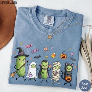 A blue t-shirt with a Halloween-themed design featuring a group of anthropomorphic vegetables dressed in costumes, including a witch, ghost, pumpkin, and cat.