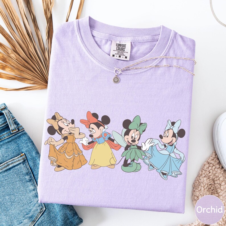a purple t-shirt with a graphic of four Disney Minnie Mouse characters, including Snow White, Cinderella, Minnie Mouse, and Mickey Mouse, standing together.