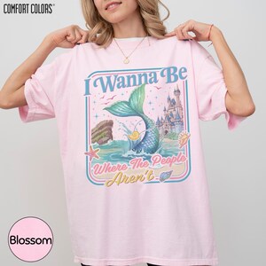 A woman wearing a pink t-shirt with a graphic design featuring a mermaid, castle, and text.