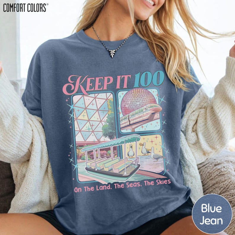 A woman wearing a blue t-shirt with a graphic design featuring a pink and blue spaceship, a boat, and a beach scene.