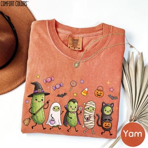 a t-shirt with a Halloween-themed design, including a group of cartoon vegetables dressed in costumes and surrounded by Halloween-related elements such as bats, pumpkins, and candy canes.
