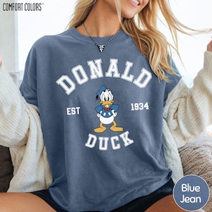 A woman wearing a blue t-shirt with a graphic of a cartoon duck and text that says "DONALD EST. 1934 DUCK".