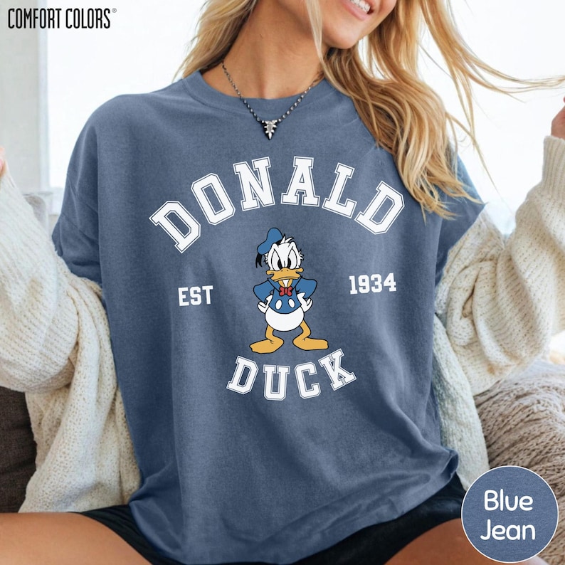 A woman wearing a blue t-shirt with a graphic of a cartoon duck and text that says "DONALD EST. 1934 DUCK".