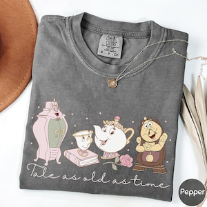 Peut inclure: Un t-shirt gris Comfort Colors avec un motif de dessin animé de La Belle et la Bête. Le motif comprend des personnages, une tasse à thé, une théière et le texte "Tale as old as time". Un collier argenté avec un pendentif est drapé sur le t-shirt.