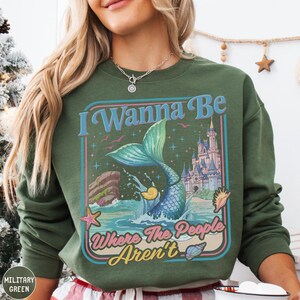 A woman wearing a green sweatshirt with a colorful graphic design featuring a mermaid, castle, and text.
