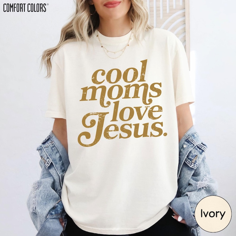 A woman wearing a white t-shirt with the text "cool moms love Jesus" printed on it, along with a denim jacket.