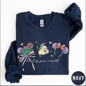 A navy blue sweatshirt with a colorful graphic design featuring a cartoon fish and flowers, along with the text "Part of your world."
