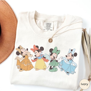 A white t-shirt with a graphic of four Disney Minnie Mouse characters in different outfits, along with a necklace and a brown hat.