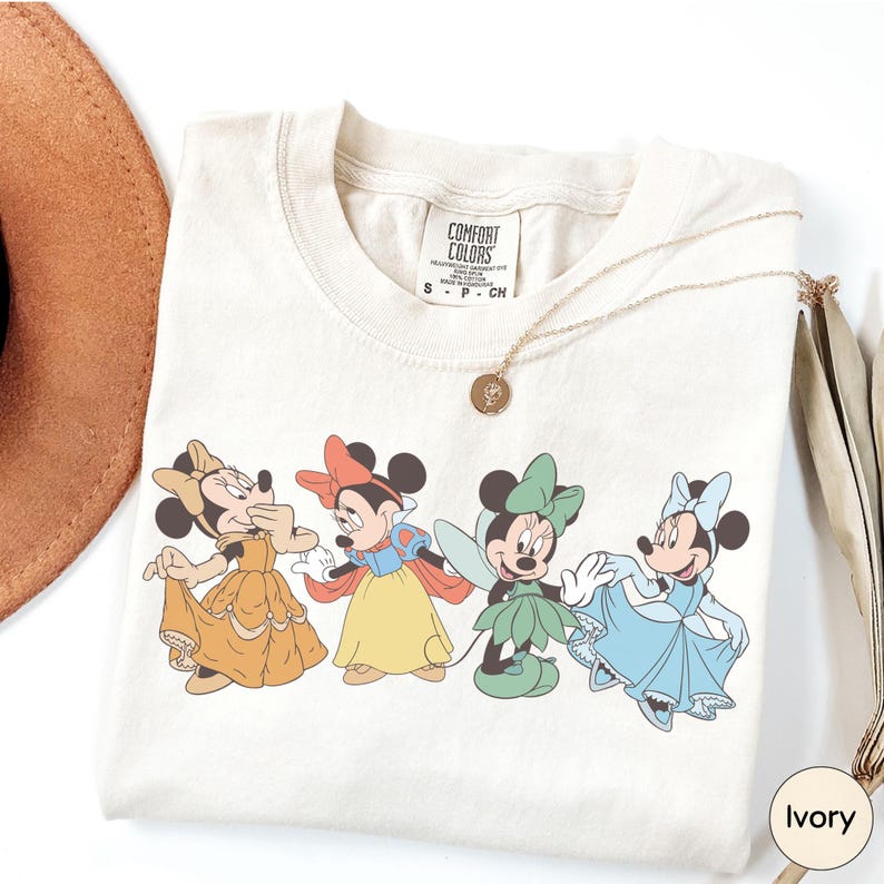 A white t-shirt with a graphic of four Disney Minnie Mouse characters in different outfits, along with a necklace and a brown hat.