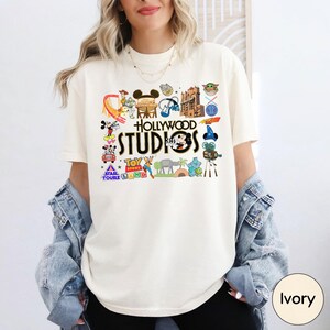 A woman wearing a white t-shirt with various Disney and Hollywood-themed graphics and text, including Mickey Mouse, the Walt Disney World logo, and the words "Hollywood STUDIOS".