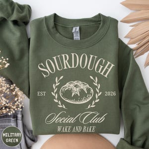 May include: A military green sweatshirt with the text "SOURDOUGH Social Club" in a white, decorative font. The design includes a bread illustration, the text "EST 2026", and "WAKE AND BAKE". The sweatshirt is a comfortable, casual style.