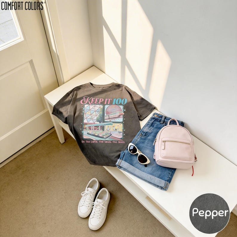 a casual outfit consisting of a black t-shirt with a graphic design, a pair of blue jeans, white sneakers, and a pink backpack, all laid out on a white surface.