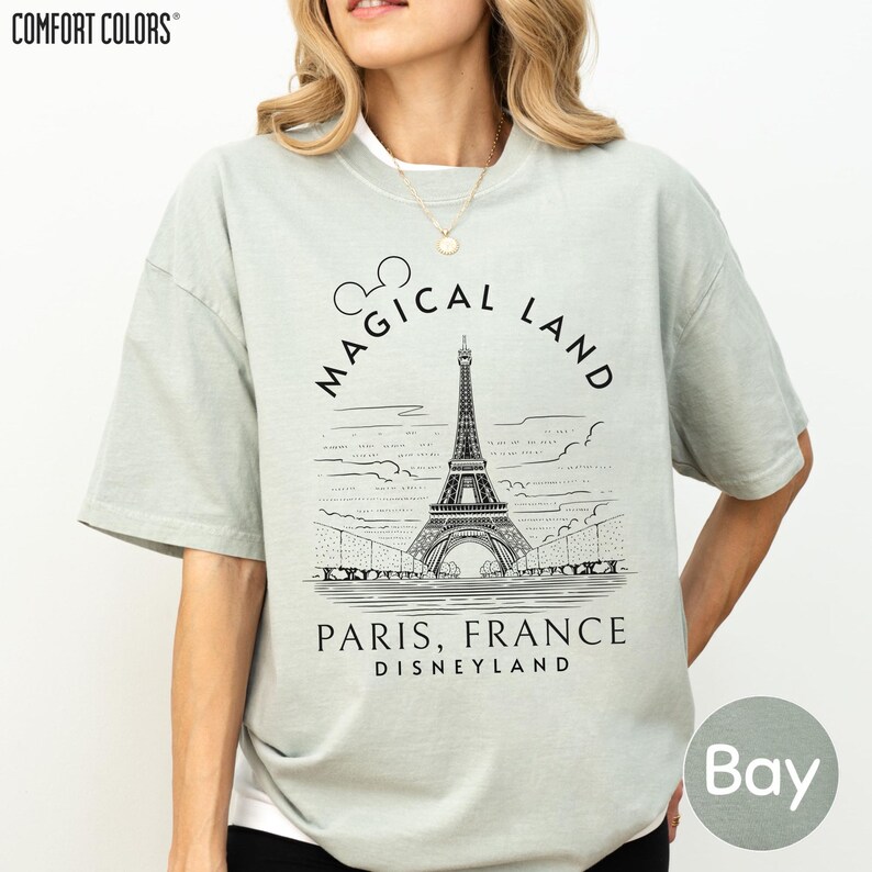 A woman wearing a gray t-shirt with a graphic design featuring the Eiffel Tower and text that says "Magical Land Paris, France Disneyland".