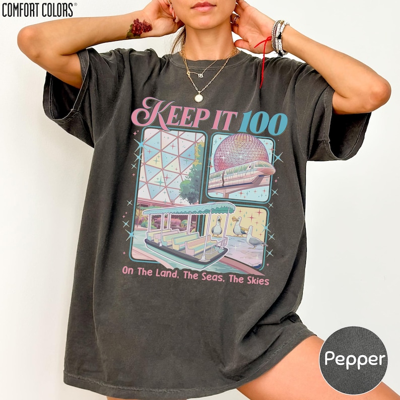 A person wearing a black t-shirt with a graphic design depicting a futuristic cityscape and the text "Keep it 100 On the land, the seas, the skies" printed on it.