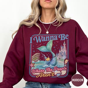 A woman wearing a burgundy sweatshirt with a colorful graphic design featuring a mermaid, a castle, and the text "I Wanna Be Where The People Aren't".