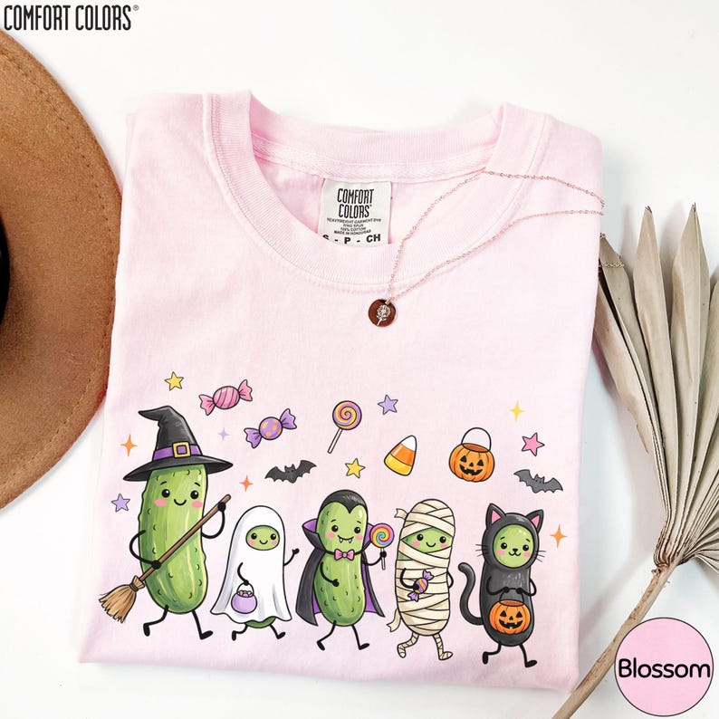 A pink t-shirt with a Halloween-themed graphic design featuring a group of cartoon characters dressed in costumes, including a witch, pumpkin, ghost, mummy, and cat.