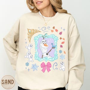A woman wearing a beige sweater with a colorful graphic design featuring characters from the Disney movie Frozen.