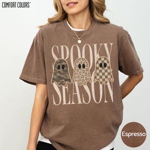 May include: A brown short-sleeve t-shirt with the text "SPOOKY SEASON" in large white letters. Three ghost illustrations are centered above the text, featuring camouflage, leopard print, and checkerboard patterns. The shirt has a crew neckline.