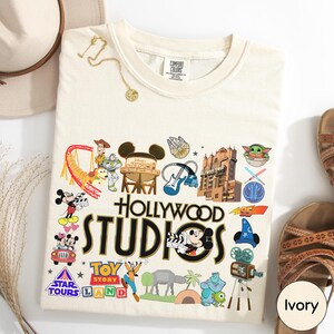 a white t-shirt with various Disney and Pixar-themed graphics and logos, including Mickey Mouse, Minnie Mouse, Toy Story, Star Tours, and the Hollywood Studios logo.