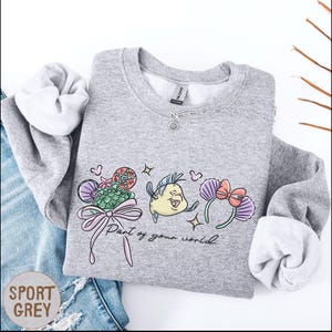 A gray sweatshirt with a cartoon character and text printed on it, along with a necklace and some jeans.