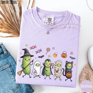a purple t-shirt with a Halloween-themed graphic design, including a witch, pumpkin, ghost, cat, and cucumber characters.