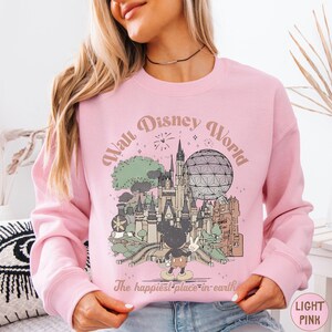 A woman wearing a pink sweatshirt with a graphic of Disney World and the text "The happiest place in earth" printed on it.