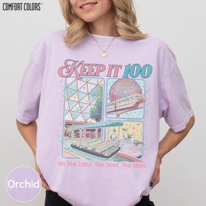 A woman wearing a pink t-shirt with a graphic design depicting a theme park ride and the text "Keep it 100 On the land, the seas, the skies".