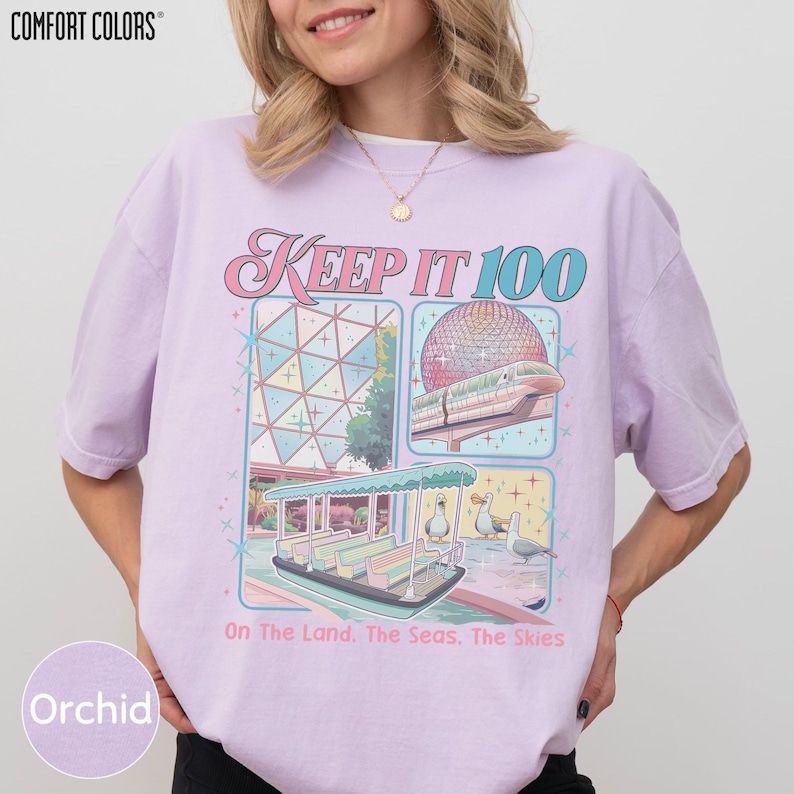 A woman wearing a pink t-shirt with a graphic design depicting a theme park ride and the text "Keep it 100 On the land, the seas, the skies".