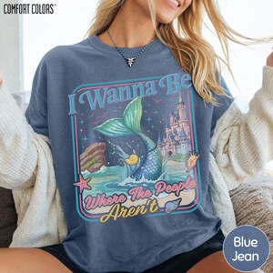 A woman wearing a blue t-shirt with a graphic design featuring a mermaid, castle, and text.