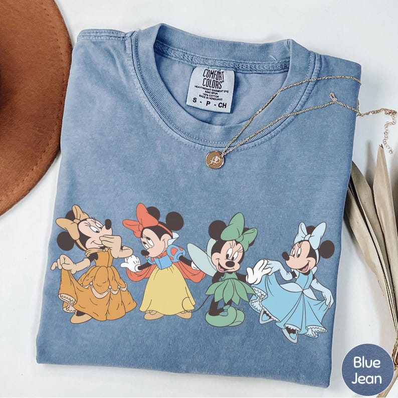 A blue t-shirt with a graphic of four Disney Minnie Mouse characters in different outfits, along with a necklace and a brown hat.