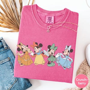 A pink t-shirt with a cartoon image of Minnie Mouse and her friends, along with a necklace and some jeans, is displayed on a white surface.