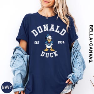 A woman wearing a navy blue t-shirt with a graphic of a cartoon duck and text that says "DONALD DUCK EST 1934". She is also wearing a denim jacket and has her hands in her pockets.