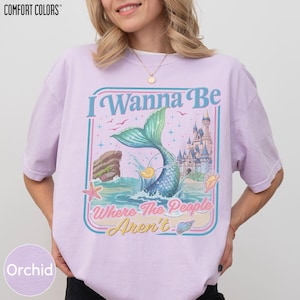 A woman wearing a pink t-shirt with a graphic design featuring a mermaid, a castle, and the text "I Wanna Be Where The People Are" and "Orchid".