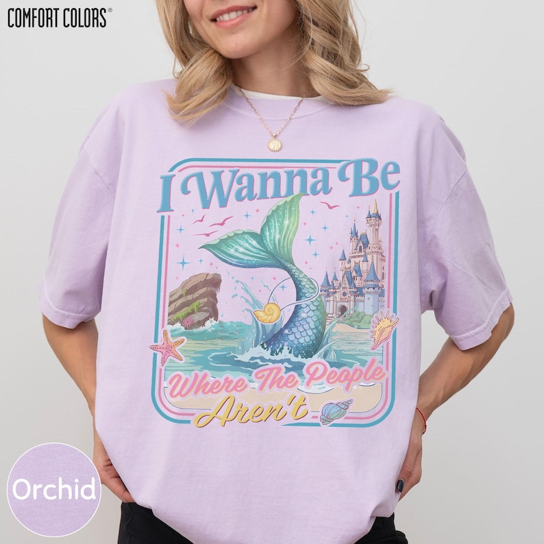 A woman wearing a pink t-shirt with a graphic design featuring a mermaid, a castle, and the text "I Wanna Be Where The People Are" and "Orchid".