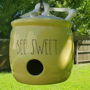 May include: A yellow ceramic bee-shaped birdhouse with a hole for birds to enter. The birdhouse has a black chain for hanging and the words "BEE SWEET" written on the front.