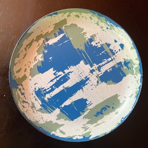 May include: A round, blue, green, and white ceramic plate with an abstract design. The plate has a distressed look, with the paint appearing to be chipped and faded.