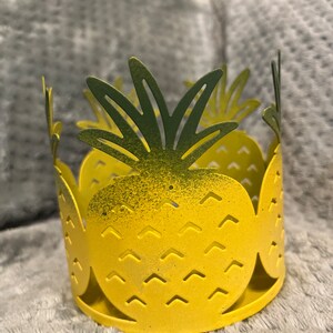 May include: Yellow metal pineapple candle holder with green leaves. The holder has a textured surface and is designed to hold a candle.
