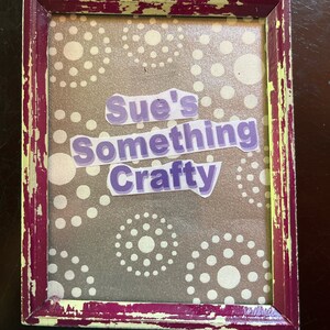 Könnte beinhalten: Ein lila-weißes Schild mit weißem Hintergrund und einem lila-farbigen Tupfenmuster. Das Schild trägt die Aufschrift "Sue's Something Crafty".