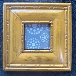 Könnte beinhalten: Quadratischer, goldfarbener Bilderrahmen mit strukturierter Oberfläche. Der Rahmen hat einen dekorativen Rand und einen blauen Hintergrund mit weißen kreisförmigen Mustern. Der Rahmen ist etwa 15 cm groß.